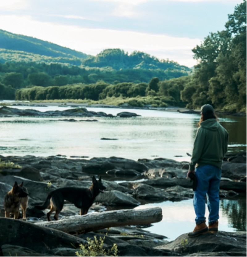 Noah Kahan with his two dogs standing by a river on rocky terrain, surrounded by lush trees and green hills.