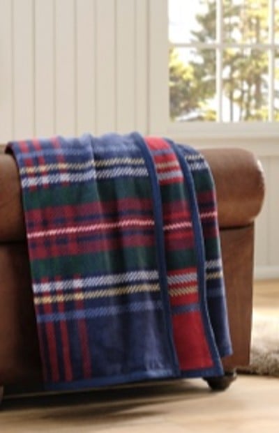 Plaid blanket with red, green, blue, and white stripes draped over brown leather chair; wood floor and window with trees outside.