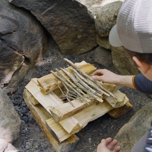 Nate placing twigs across the top of log cabin style stacked wood to form a roof.