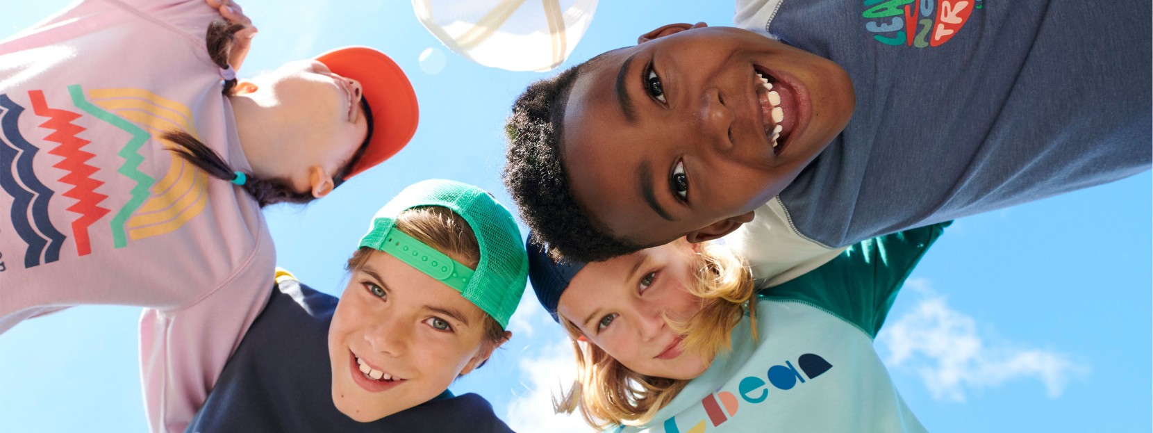 4 smiling kids in a huddle, shot from below looking up at faces and blue sky.
