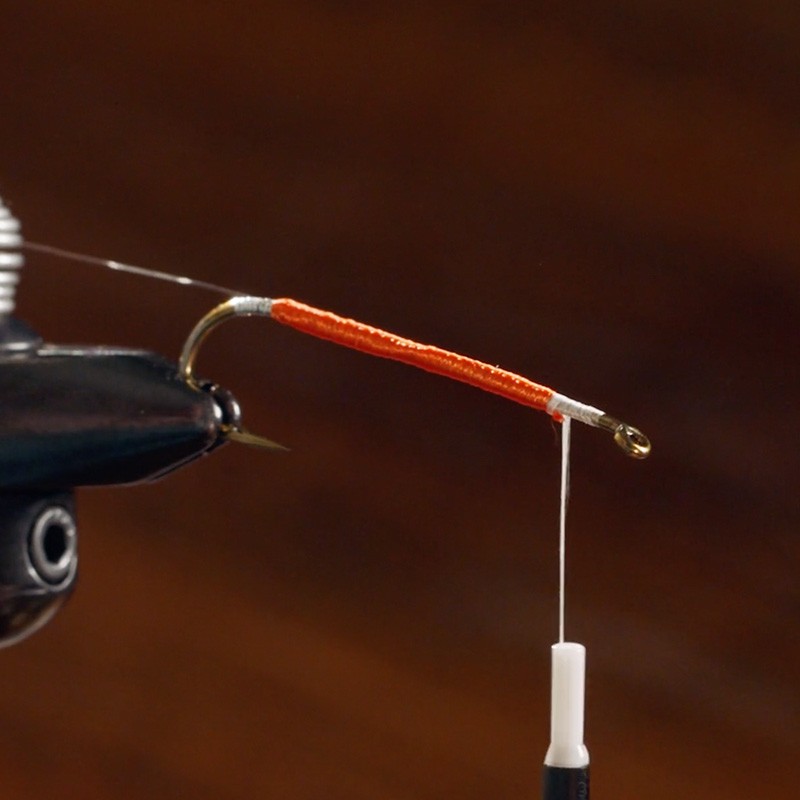 Close-up of a fishing hook being wrapped with orange thread, held in place by a vise.