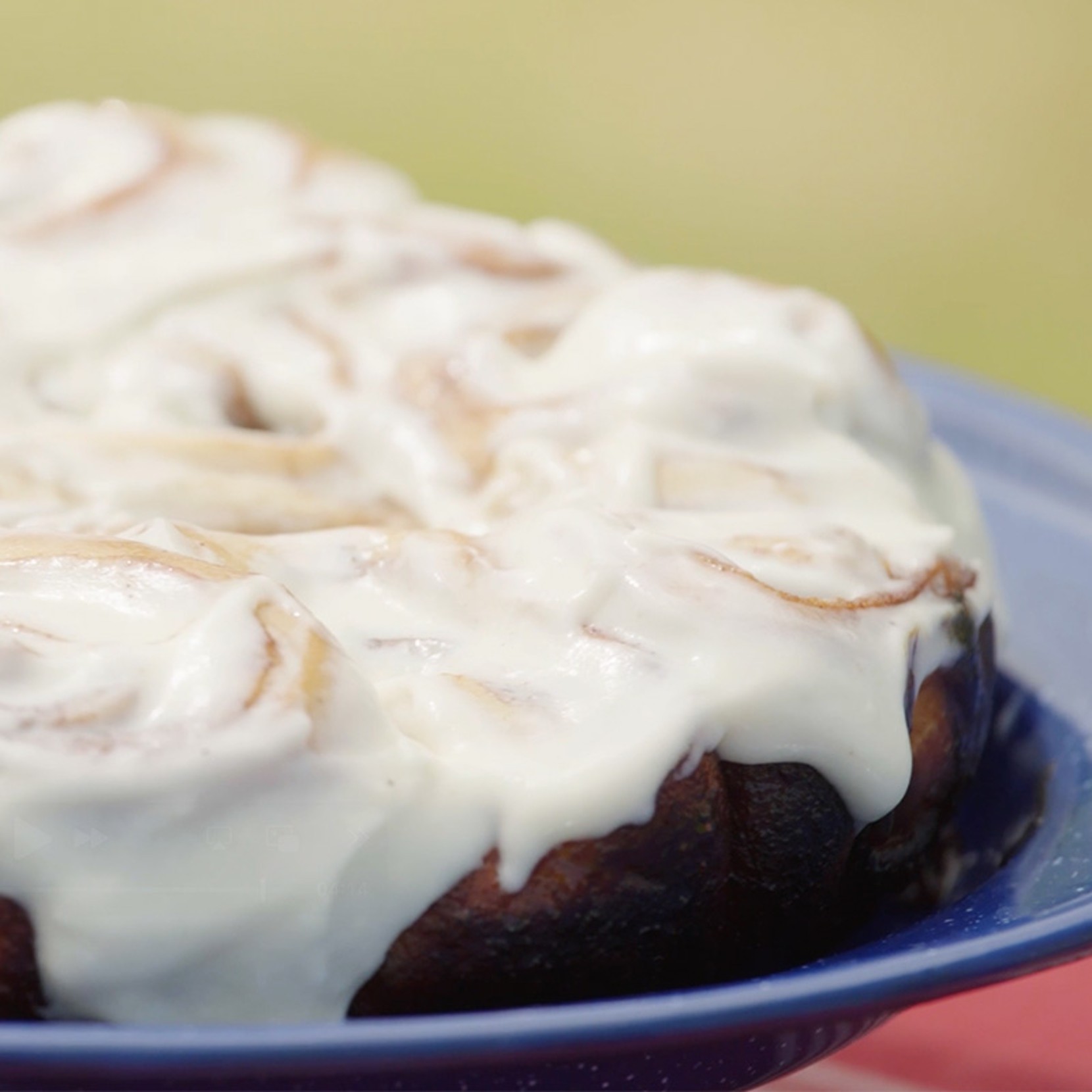 Close-up of a cinnamon roll cake with white icing on a blue plate, set against a softly blurred background.