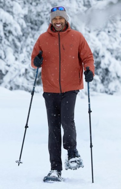 Person wearing an orange jacket, black pants, and snowshoes walks through snowy terrain using trekking poles.