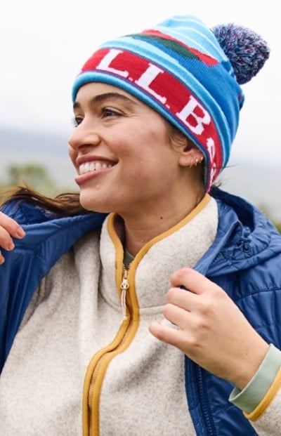 A smiling woman in blue and red beanie, white fleece with yellow zipper, and blue jacket stands outdoors.