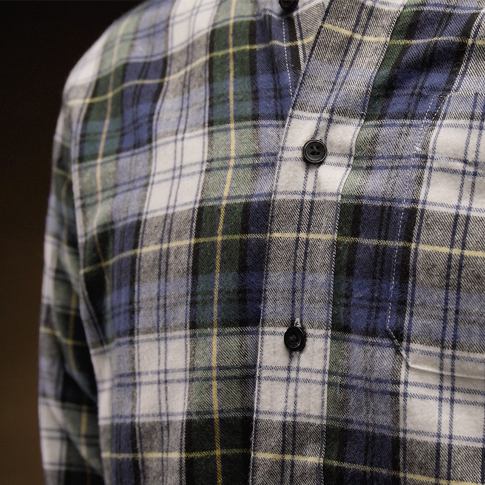 Close-up of a plaid shirt with blue, green, white, and black patterns, featuring black buttons against a dark background.