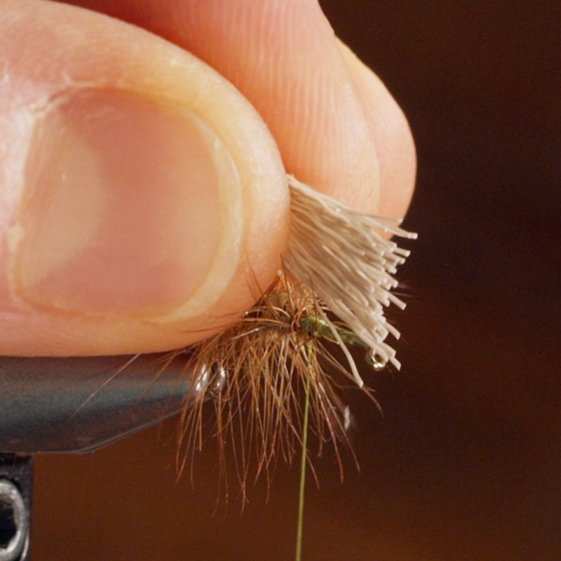 A hand holding a bundle of fibers, attaching them to a fishing fly with brown and green materials.