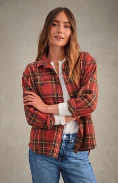 Woman in red plaid shirt over white top and blue jeans stands with arms crossed against a plain background.