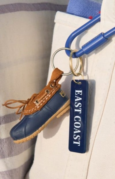 Mini bootā€‘shaped keychain and an “EAST COAST” tag hanging from a blue carabiner clipped to light-colored canvas bag.