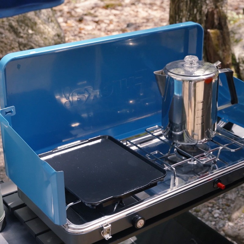 A portable stove with a coffee percolator on it, set outdoors, suggesting camping or a picnic. 