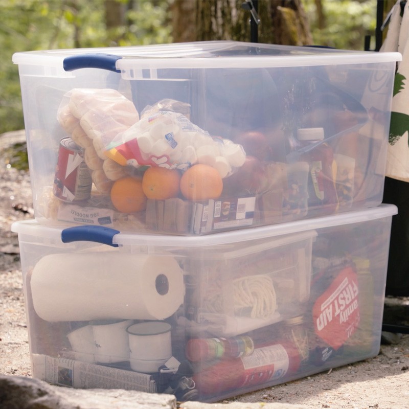 Two stacked transparent storage bins filled with various supplies, labeled “FIRST AID,” in a forest setting.
