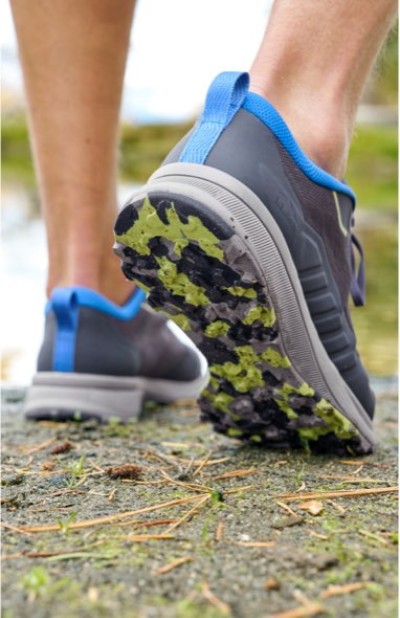 Gray trail running shoes with blue accents and green-tread soles, shown mid-step on a dirt path with scattered pine needles.