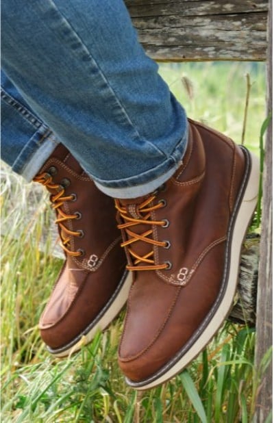 Close up of brown leather boots with yellow laces resting on a wooden fence above green grass.