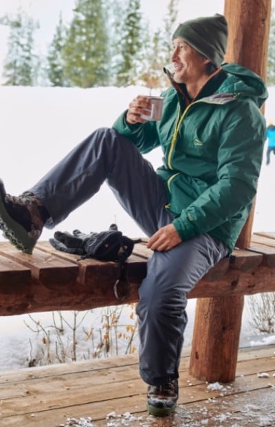 A man in outdoor winter clothing sits on a wooden bench, holding a cup, with snowy scenery in the background.
