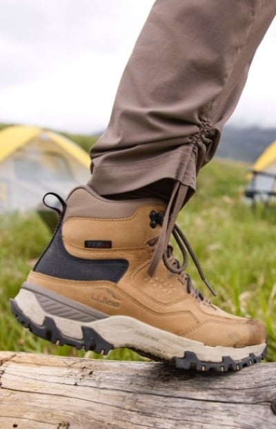 Person in brown hiking boots and pants steps on a log in a grassy outdoor area with tents visible in the background.