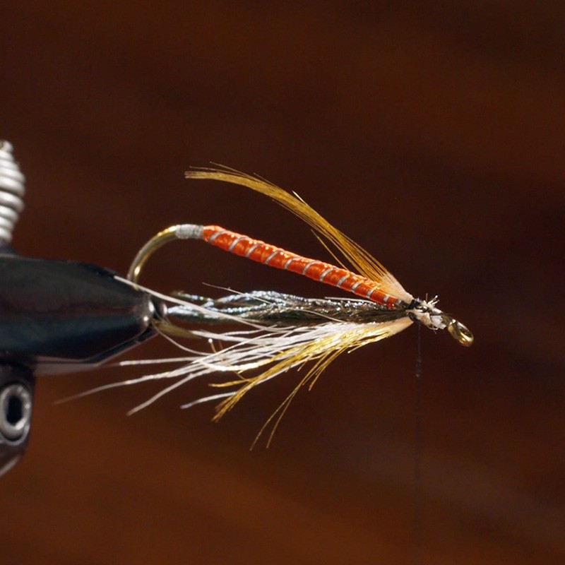 Copilot Close-up of a detailed, handmade fishing fly with an orange body, gold hook, and white and yellow feather accents.