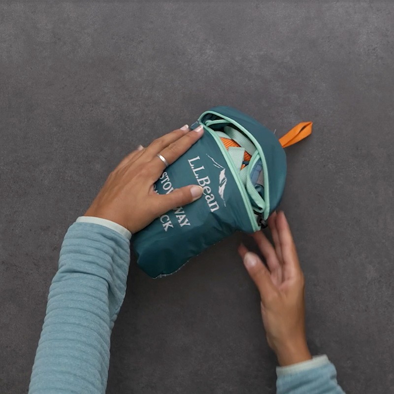 Hands fold the zipper flap of a teal Stowaway Pack from the back of the pocket over to the front.
