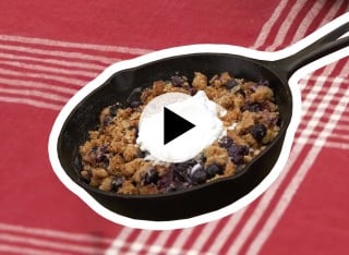A skillet with blueberry cobbler topped with a dollop of whipped cream on a tablecloth with a play video icon in the center.