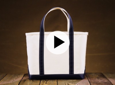 Beige canvas tote with dark blue handles and trim, on wood surface with brown backdrop and play button overlay.