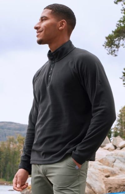 Person wearing a dark long‑sleeve pullover and light pants, standing outdoors near rocks and trees.