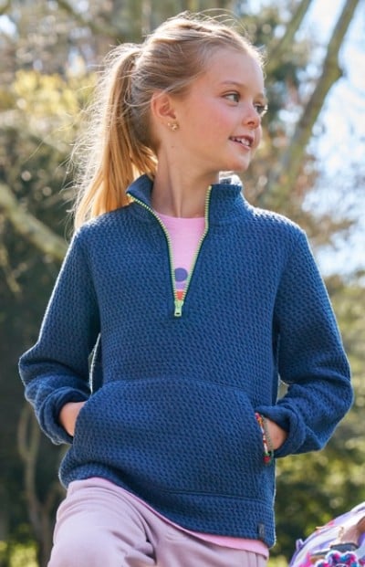 A girl in a blue pullover with front pocket stands outdoors among trees, sunlight filtering through.