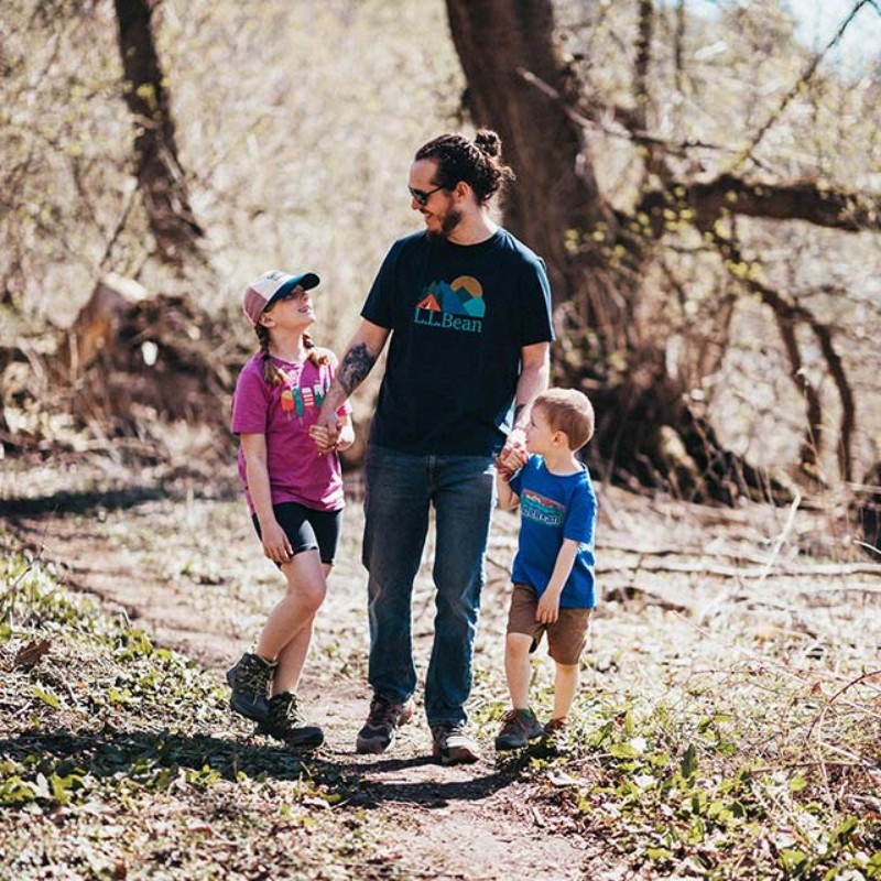 A dad holding hands with two young kids while walking on a wooded trail.