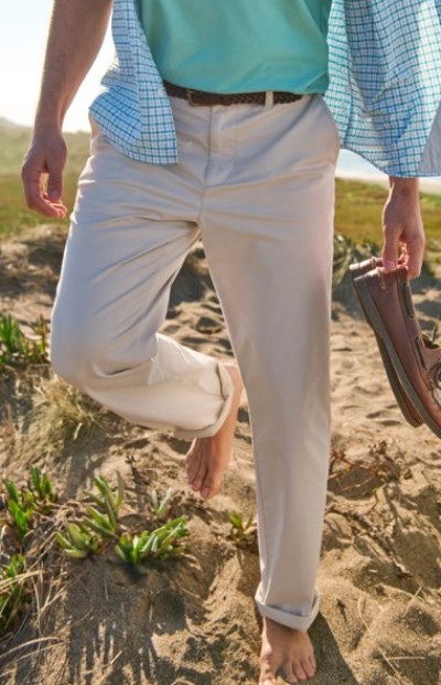 Barefoot person walking on sand wearing light pants, a teal shirt, and a checkered button‑up, holding a pair of shoes.