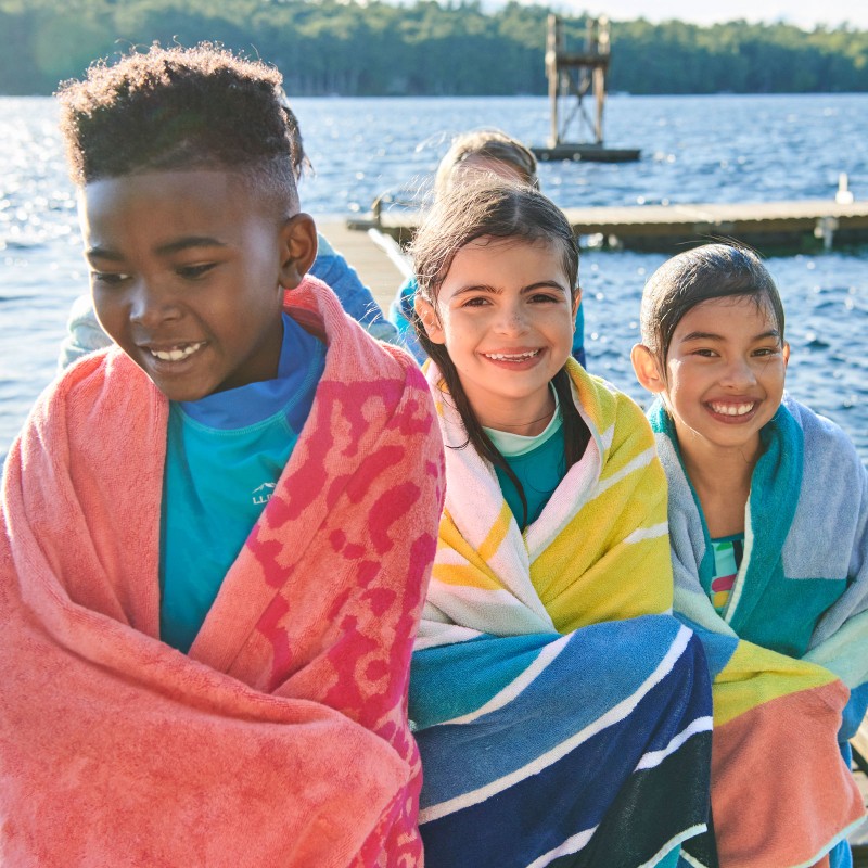 Smiling kids wrapped in beach towels by a dock on a lake.