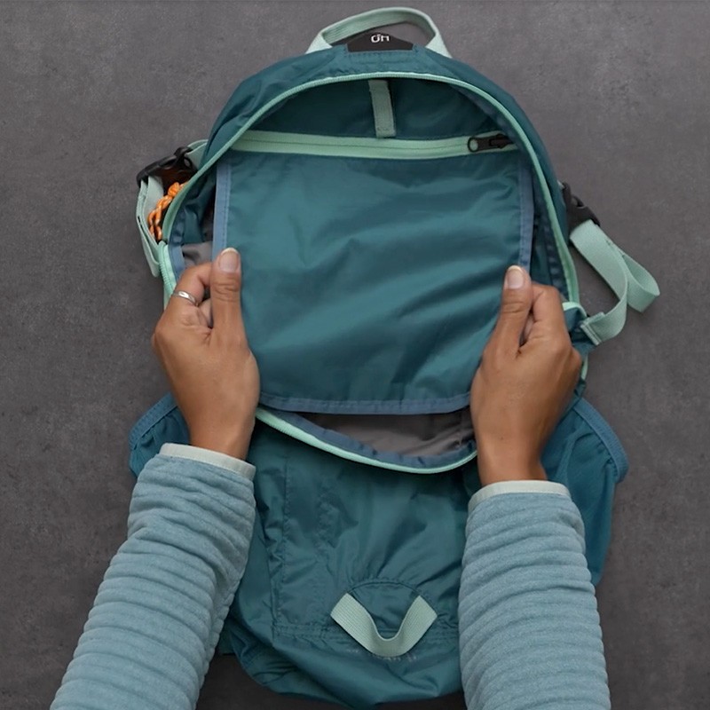 Hands open a teal Stowaway Pack revealing it's zippered interior pocket.