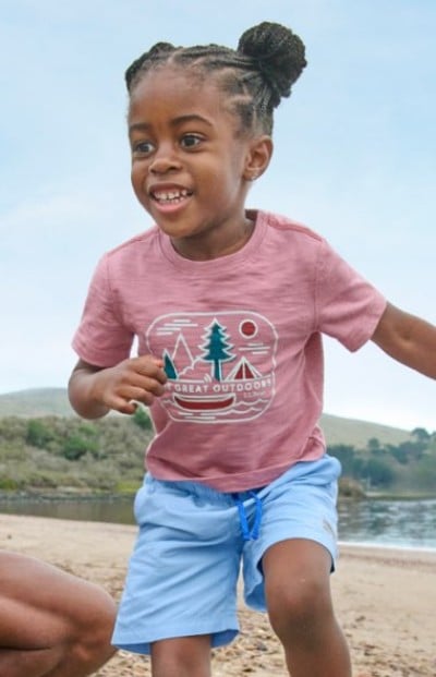 Child running on a sandy beach, wearing a pink graphic T‑shirt and light blue shorts with water and hills in the background.