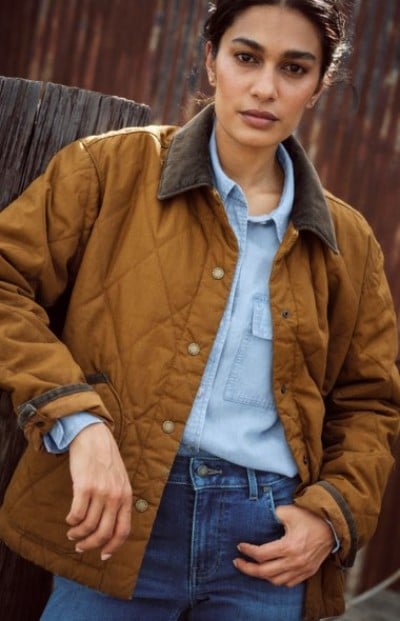 Person wearing a brown jacket layered over a denim shirt in a rustic setting.