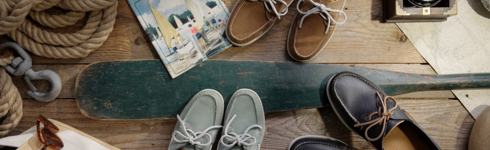Vintage nautical-themed display with several pairs of boat shoes, an oar, maps, catalog, and sunglasses on wooden planks.