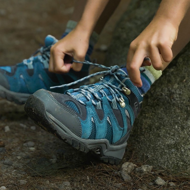 A child tying the laces of a blue hiking boot while sitting on a rock.