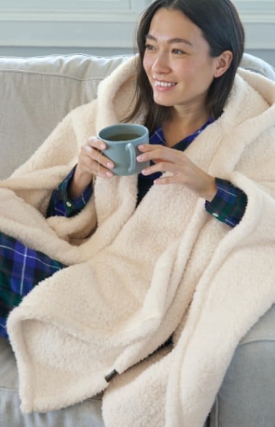 A woman wrapped in a cream colored throw sits on a boat smiling while holding a mug of coffee.