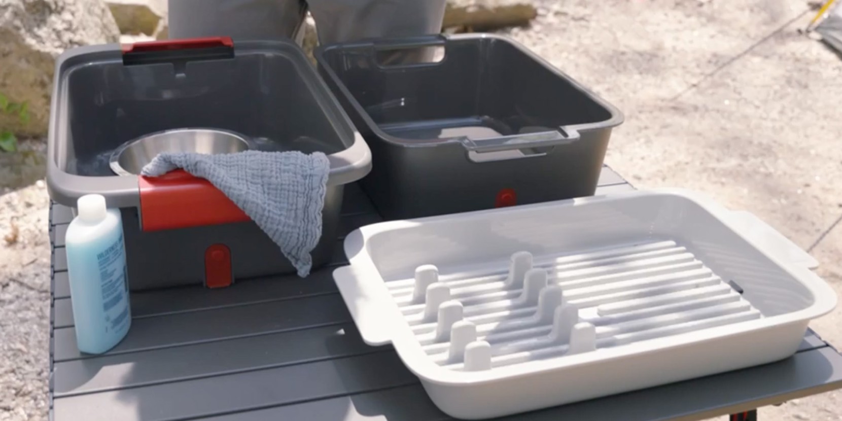 A person is washing dishes outdoors using two bins and a drying rack on a table, suggesting a camping or picnic cleanup.