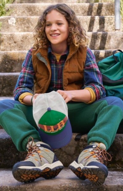 Child in plaid shirt, brown vest, green pants, and hiking boots sits on stone steps holding a cap labeled L. L. B.