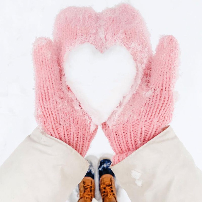 A person with pink mittens holding snow in the shape of a heart.
