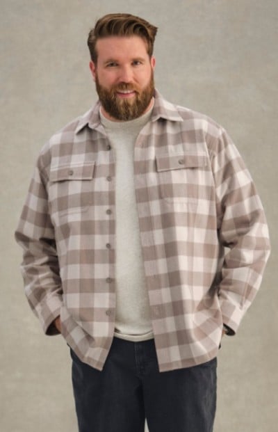 Man in beige and white plaid shirt over light gray t-shirt stands with hands in pockets against a plain background.