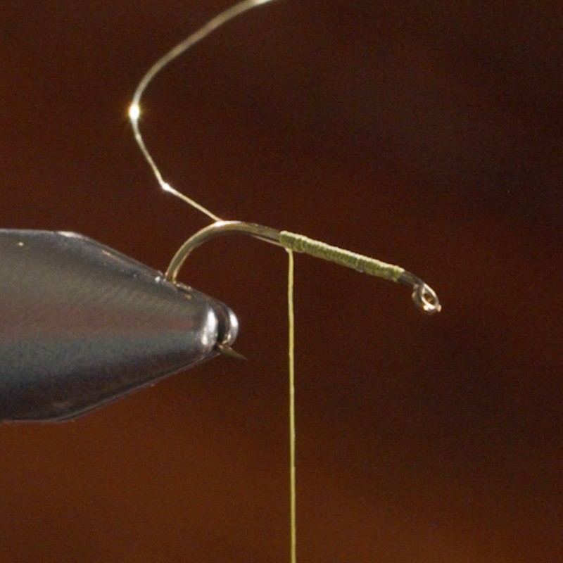 A close-up of a fly-tying vise holding a hook with green thread wrapped around the shank, used for making fishing flies.