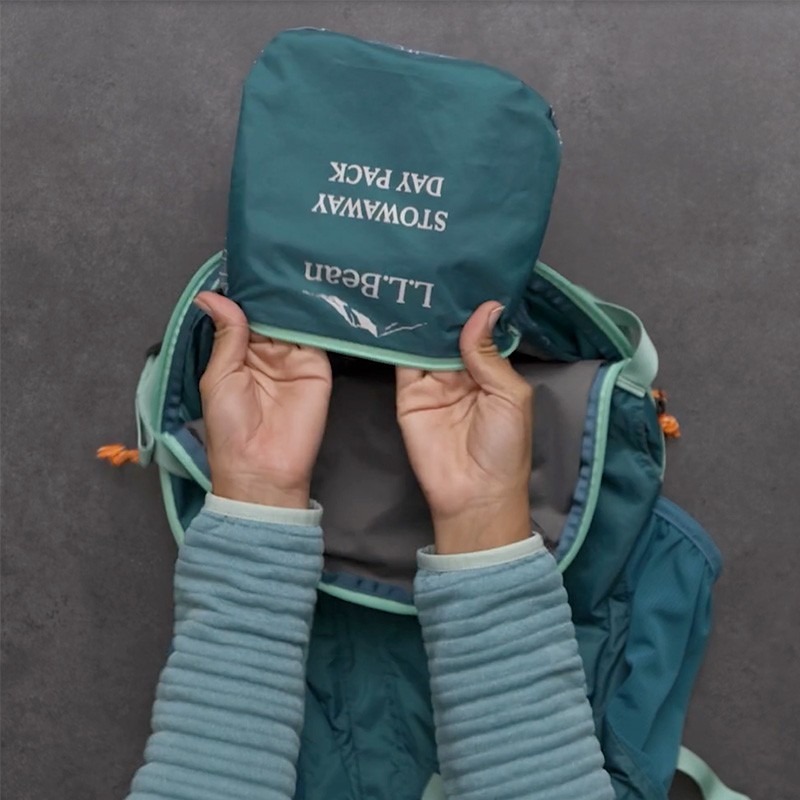 Hands pull the teal Stowaway Pack's interior pocket inside out revealing the L L Bean logo and pack name.