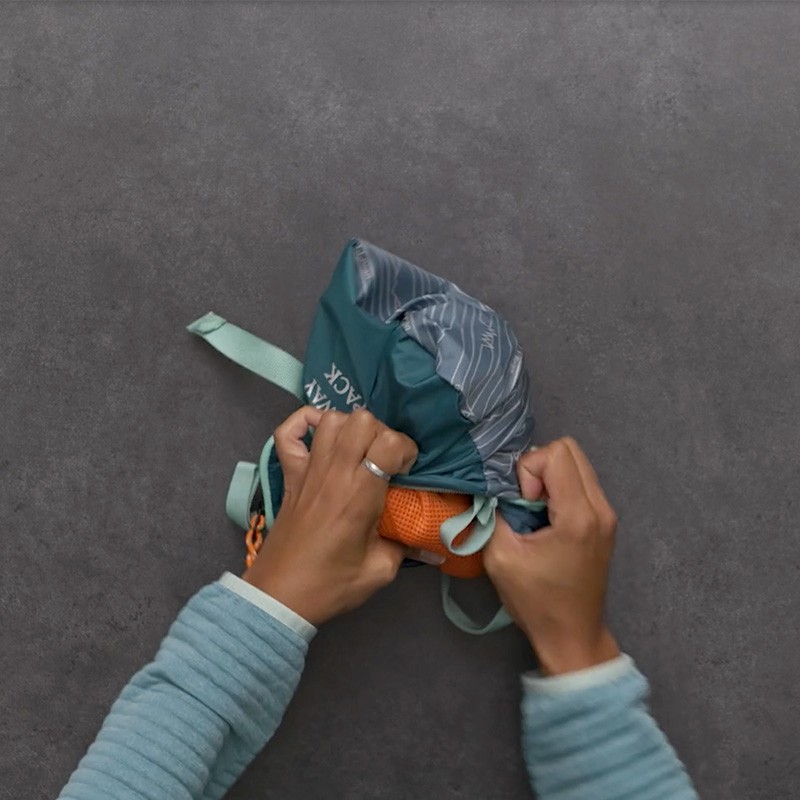 A teal Stowaway Pack is turned over as hands stuff the pack into it's inner pocket.