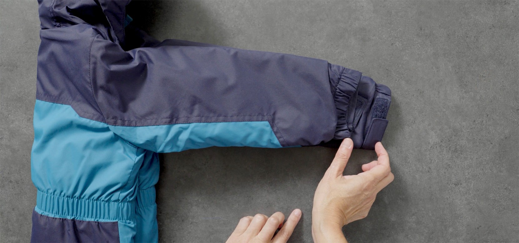 Hands adjust the sleeve of a blue and navy winter jacket on a gray surface.