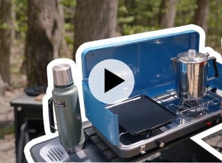 A portable stove with a coffee percolator on it, set outdoors. A play video icon is in the center.