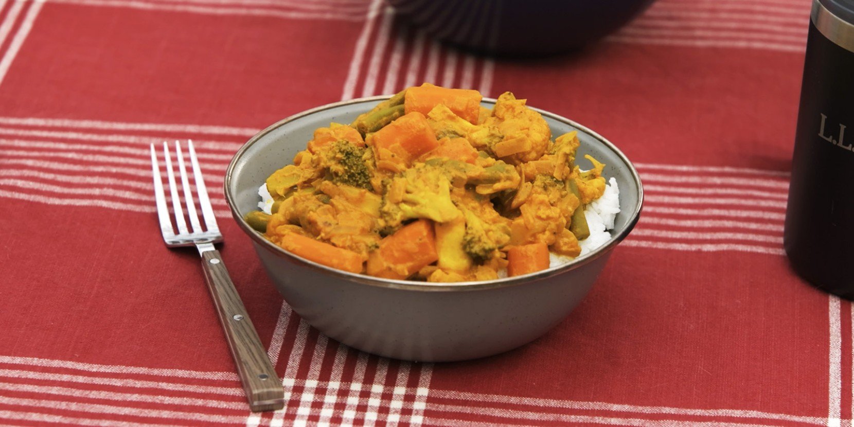 A bowl of vegetable curry on rice sits on a red and white checkered tablecloth.