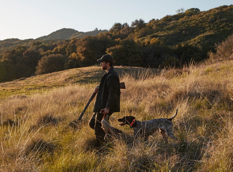 A hunter and his dog walking through a grassland.