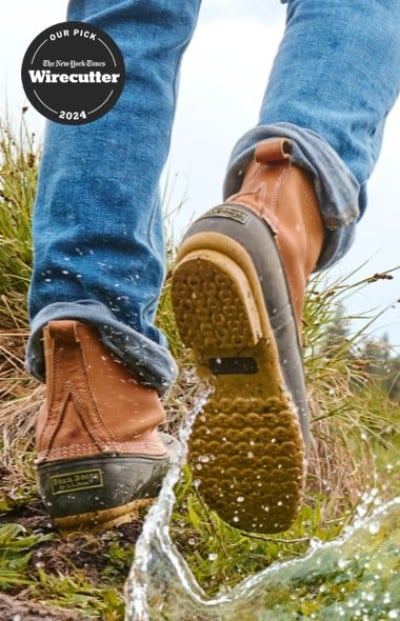 Close-up of Bean Boots splashing through water on a grassy trail and labeled as Wirecutter’s 2024 pick.