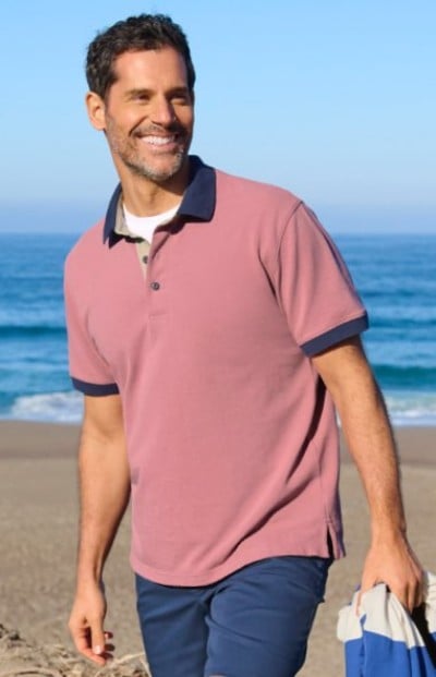 Man wearing a pink polo with dark trim, holding clothing while walking on a beach near the ocean.