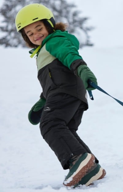 Child in green snowsuit, yellow helmet, and green gloves stands on snow holding a blue strap, wearing boots with brown soles.