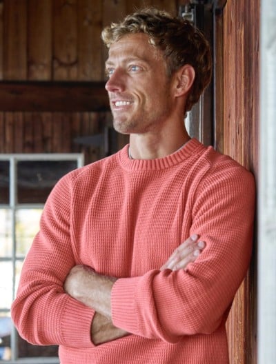 Man in a coral sweater stands with arms crossed against a wooden wall. 