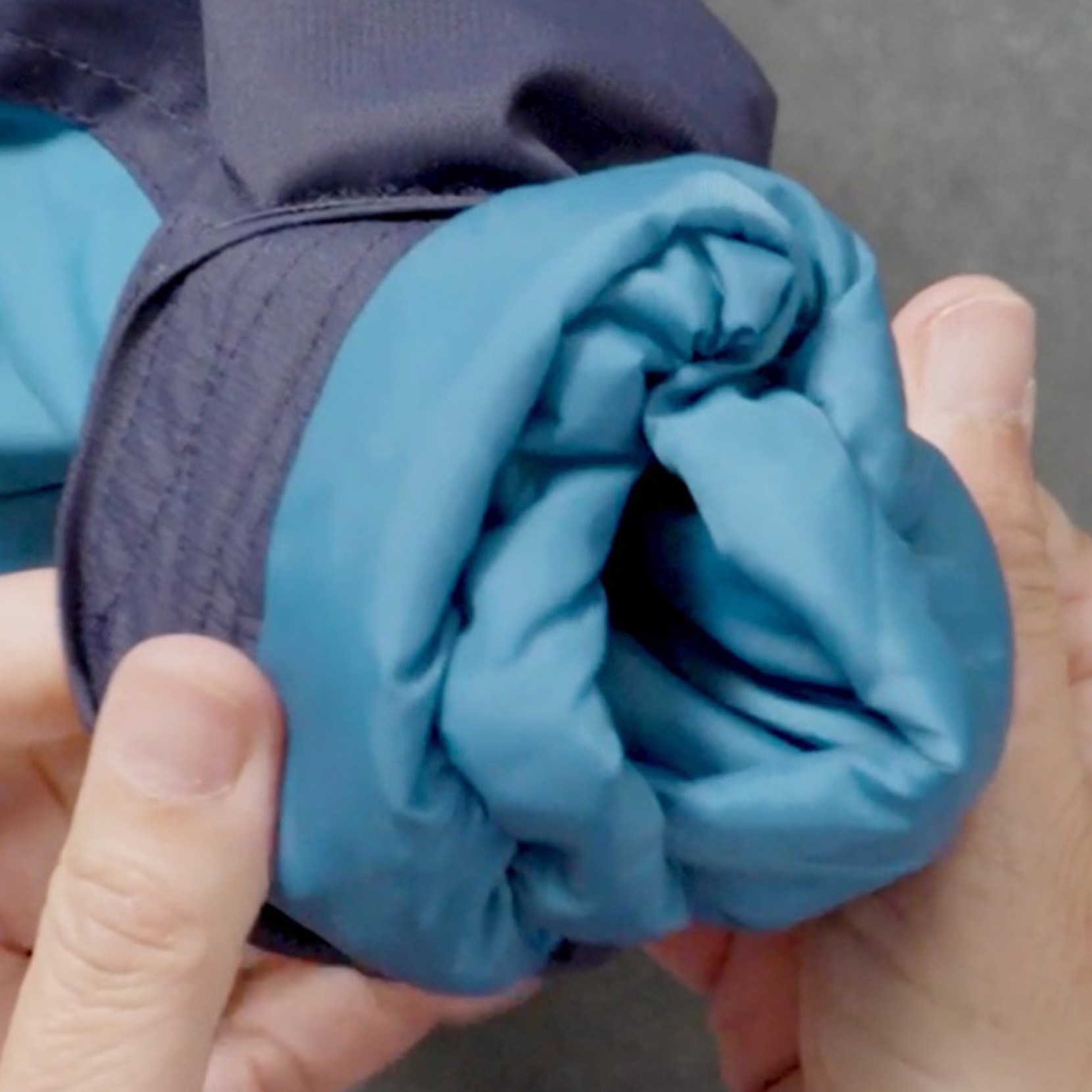 Two hands hold the rolled-up sleeve of a blue and teal jacket against a plain gray background.