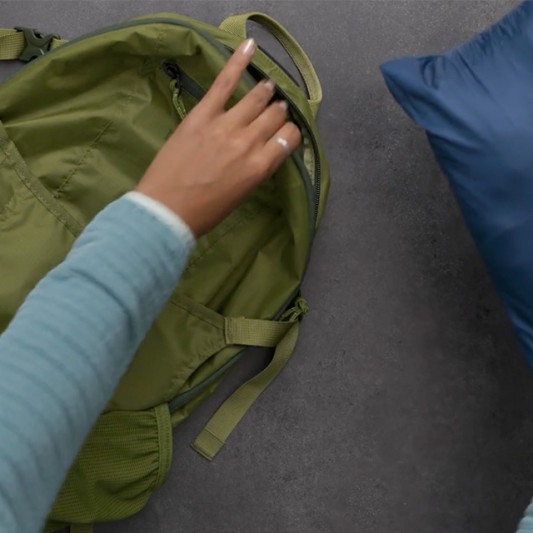 A hand opens a green backpack with a blue stowawayed jacket resting nearby.